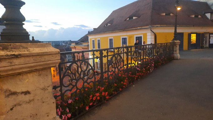 An outdoor evening scene features a yellow building with a red roof behind an ornate black metal railing decorated with pink flowers.