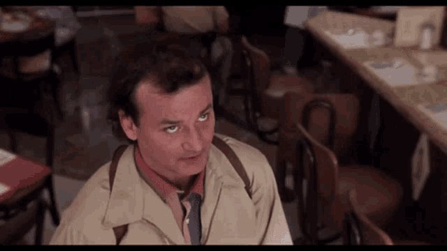 The image features a man sitting at a table with his arms crossed, wearing a tan jacket and tie. The background shows multiple chairs and dining tables arranged around the room. There are also other people present in the scene, but they are not the main focus of the image. The caption "Happy Father's Day cap also the wait is unbearable WEN APEFEST?!" seems to be related to a post on social media about waiting for food at a restaurant or a similar situation.

The man sitting at the table appears to be looking up, possibly observing something happening in the room or engaging with someone nearby. The image captures a moment of restlessness and frustration due to the wait time, which could be related to the caption provided by the post.