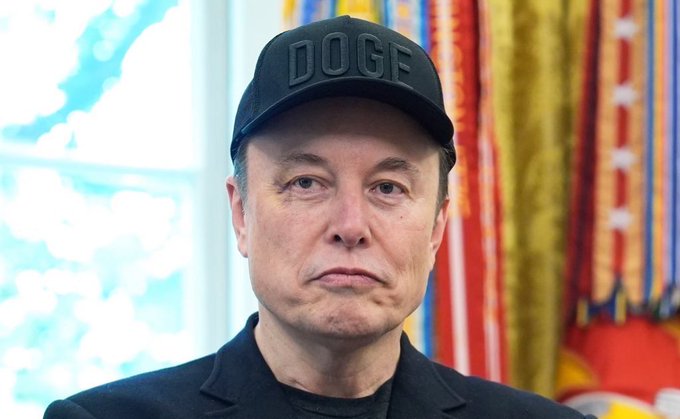 A photograph of Elon Musk wearing a black baseball cap with the word 'DOGE' embossed on it, a black jacket, and a black shirt, standing indoors with an American flag visible in the background.