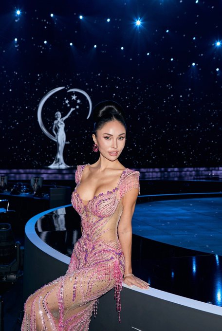 A Miss Universe Malaysia finalist poses in a beaded pink gown on stage beneath the pageant's iconic moon-and-stars backdrop, reflecting her dedication to the competition. The glamorous image contrasts with her personal dilemma of choosing between the prestigious pageant and pursuing crypto pumps in the bull market.