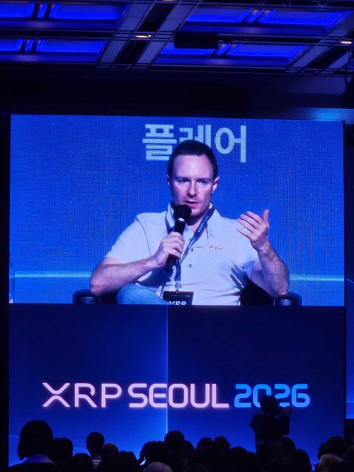 A man speaks into a microphone on a large screen at the "XRP SEOUL 2026" conference, with Korean text "플레어" (Flare) displayed behind him. The image illustrates the referenced speaker slot, likely featuring Hugo Philion, as the audience watches from below at the Seoul event.
