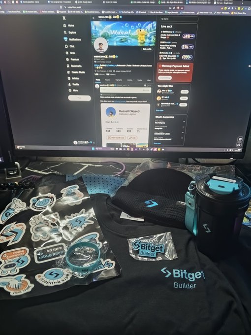The image shows a computer monitor displaying a social media profile, with a black shirt and blue hat placed next to it. There are also several stickers scattered around the desk area. The main focus of the image is the computer screen showing the person's Twitter account, which has over 100 followers.