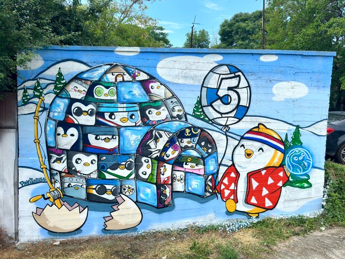 A colorful outdoor mural depicts cartoon penguins, a volleyball, and a yellow bird character on a blue background, with trees and a parking lot visible around it.