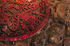 A weathered red manhole cover embossed with "2022" sits embedded in cobblestone, its warm crimson tones evoking the lasting impact of Azuki's 2022 launch—a quiet relic marking where the story began and the feeling endures.