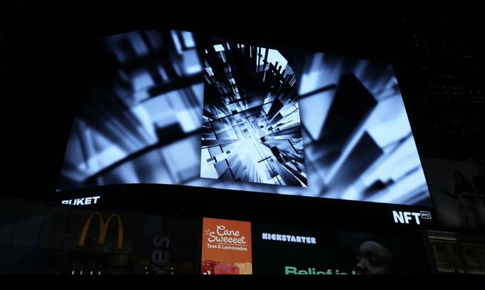 A nighttime photograph of a large illuminated billboard displaying an abstract geometric blue and white pattern, with smaller illuminated advertisements for McDonald's, Kickstarter, and NFT visible below it.