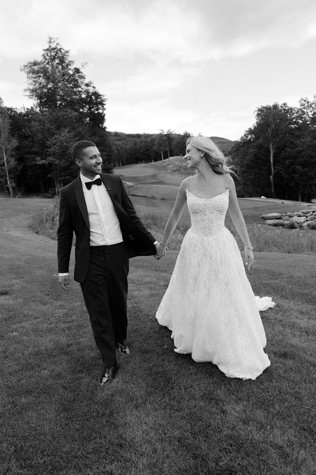 A black-and-white wedding photo shows a groom in a tuxedo and a bride in a white gown holding hands and walking on a grassy lawn.