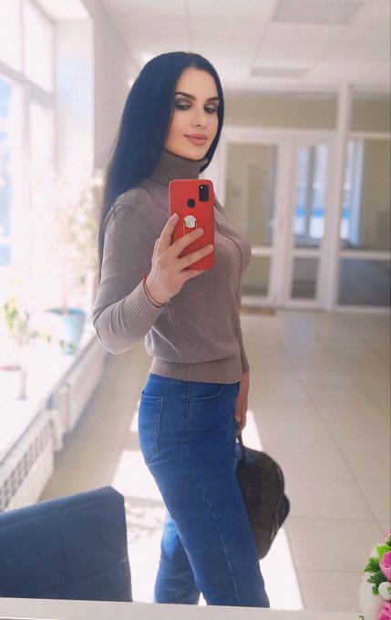 A woman with long dark hair takes a mirror selfie with a red iPhone, wearing a beige turtleneck sweater and blue jeans.