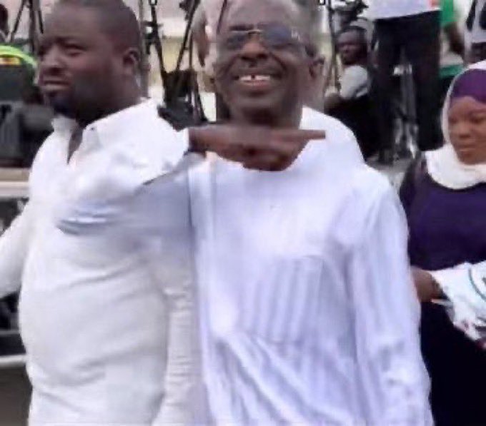 A man in white traditional attire and glasses smiles while pointing confidently at something off-camera, with another man and a woman in a headscarf standing nearby, capturing a celebratory and optimistic moment that mirrors the hopeful, meme-like anticipation of an impending crypto rally referenced in the post.