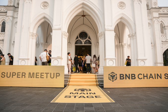The entrance to a 'BNB CHAIN SUPER MEETUP' event featuring a 'MAIN STAGE' banner on the steps and attendees gathered near the arched doorway of a white building.