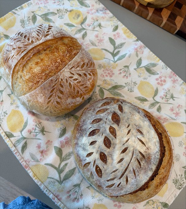 Two beautifully scored sourdough loaves—one with a wheat-ear design and the other with a leafy wreath pattern—rest on a floral lemon-print cloth, with the decorative bread scoring evoking the "bear market" theme as a playful, bread-making parallel to financial trends.