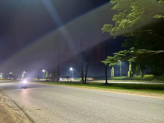 The image shows an empty street at night with a car driving down it, possibly heading towards a park or another destination. The scene has a dark background, which contrasts with the illuminated streetlights that cast light onto the road. There are no people visible in the photo, and the car is moving away from the camera's perspective.
