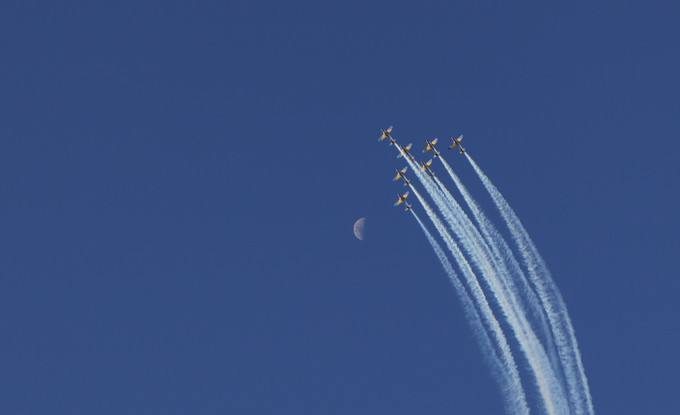 Five yellow and blue aerobatic aircraft flying in formation, leaving white smoke trails as they descend past a visible moon against a deep blue sky.