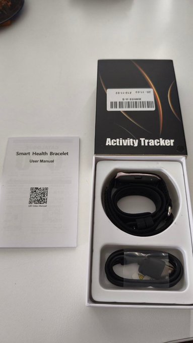 The image shows a box containing an activity tracker, possibly a smart bracelet or fitness band, with its packaging open. The box has a white background that contrasts with the black label and instructions inside. There are two main parts of the box visible - one on top and another below it. The bottom part features a USB cable for connecting to devices like smartphones or tablets. The image also includes a watermark in the bottom right corner, which could be an indication of the source or creator of the content.