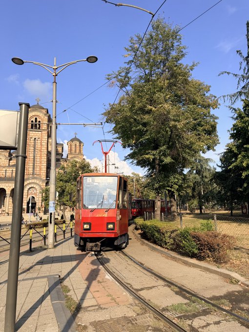A red tourist tram travels along tracks in front of a large stone church with crosses on its towers, surrounded by tall trees under a blue sky.