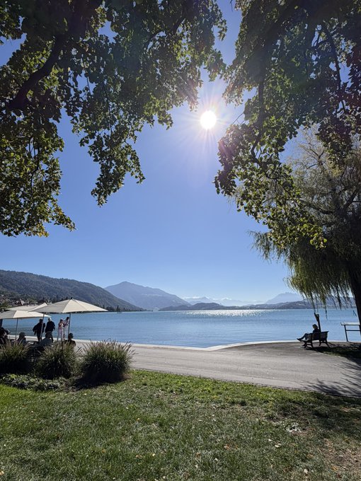 Sun shining through lush green trees over a calm lake with distant mountains, as people relax along the shore in Zug, Switzerland.