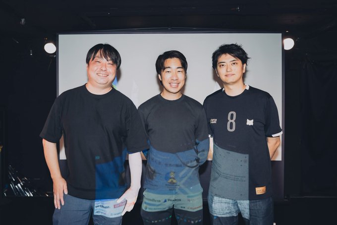 Three men stand smiling on a stage in front of a projection screen, with the center figure wearing a dark t-shirt featuring a logo and the rightmost man in a black shirt displaying the number "8" and a small cat emblem. The post identifies this as the "Enterprise Vision Summit 2026 Summer" focused on crypto market trends, with the central figure likely being Shota Watanabe (@WatanabeSota), one of the speakers at the event.