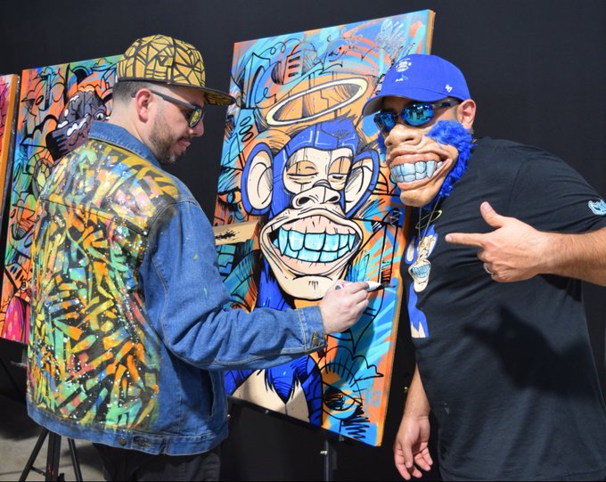 Two men pose next to a vibrant street art painting of a blue monkey with a toothy grin, one signing the artwork.
