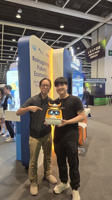 Two men pose at what appears to be a convention booth displaying "Reimagining Future Economies" signage, proudly holding a Floki-branded B.Duck Minibot mascot trophy. The image captures their meeting at the Animoca Brands booth, as referenced in the post about meeting Yat Siu to showcase their FLOKI and B.Duck Minibot robotics ecosystem.