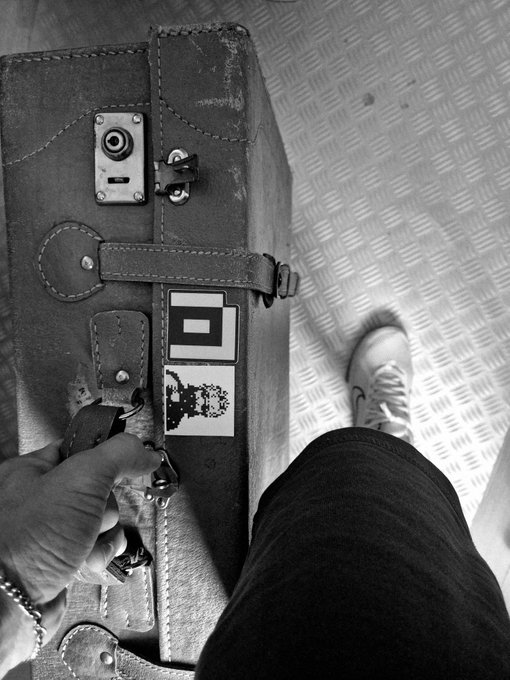 A black and white photo showing a person holding a vintage leather suitcase with stickers, while another person's foot in a sneaker is visible on a textured metal floor.