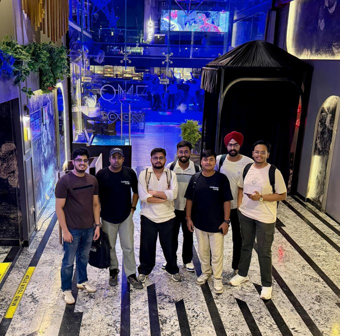 A group photo of seven men posing together inside what appears to be a modern lounge or club with blue lighting and a black canopy tent in the background.