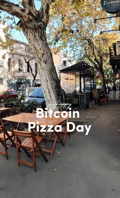 The image features a street scene with two wooden dining tables and chairs set up outside of a restaurant or cafe. The tables are surrounded by chairs, creating a welcoming outdoor seating area for customers. A banner above the tables displays "bitcoin pizza day", indicating that this is an event related to Bitcoin or cryptocurrency. The text in the image reads: "RIVER was directly used by merchants to make payments at the Argentina Pizza Festival!". This suggests that the restaurant or cafe has incorporated Bitcoin into their operations, possibly for payment purposes or as a unique way of celebrating the Bitcoin Day event.
