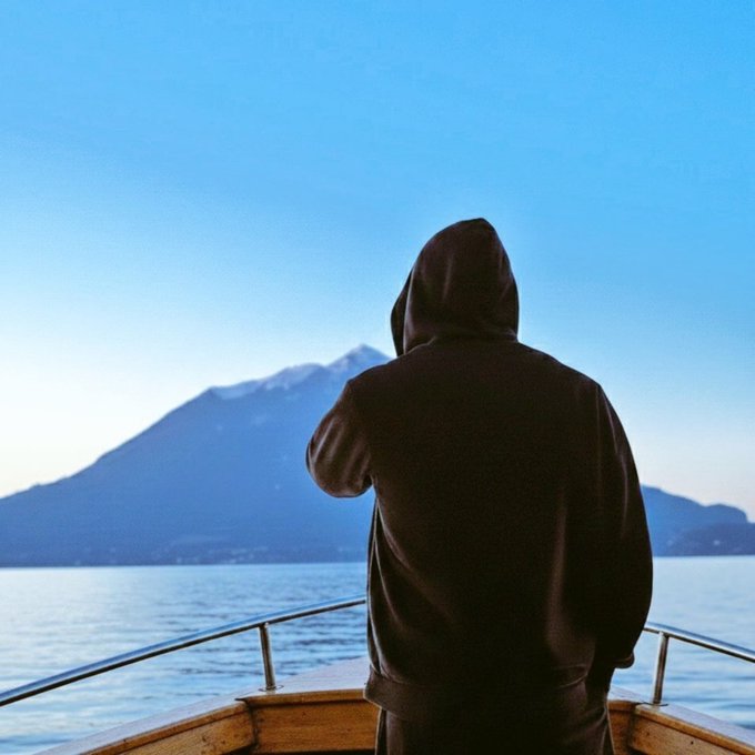 A person in a dark hooded sweatshirt stands on the bow of a boat, looking out at a large mountain rising from the sea under a clear blue sky.