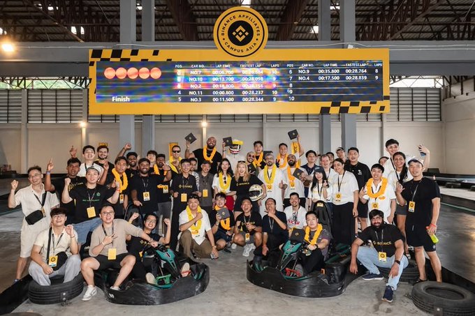 A large group of people posing with go-karts and trophies at the 'BINARY ARENA CAMPUS ROAR' event, with a digital scoreboard displaying race results above them.