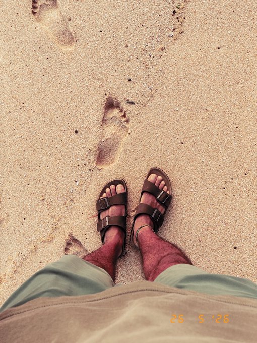 A first-person view of someone wearing sandals standing in sand, with footprints visible ahead; dated '26 5'26'.