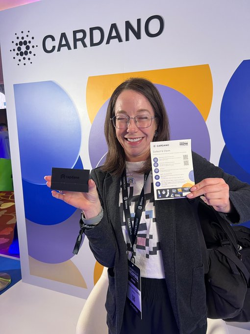 A smiling woman wearing glasses and a lanyard holds a Ledger hardware wallet and a Cardano-branded "Collect & Store" card in front of a Cardano backdrop.
