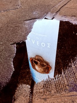 A weathered, torn cardboard box or surface featuring a faded blue label with partial Greek text 'ΑΡΚΕΟΣ' visible along with a circular metallic element.