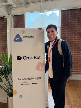 A man with a backpack stands smiling in front of a 'Welcome' sign for the 'Grok Bot Founder Build Night' event, presented by Hugging Face and MongoDB.