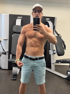 A muscular, shirtless man wearing a white cap, sunglasses, light blue shorts, and a gold chain takes a mirror selfie with his phone in a gym.