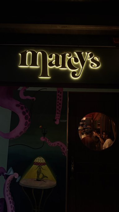 A dimly lit storefront with a glowing yellow neon sign reading 'Marcy's', featuring a round window showing people inside and octopus tentacles painted on the wall.