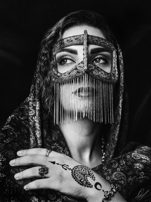 A striking black and white artistic portrait of a woman wearing an ornate beaded face veil/mask and a decorative hood, with visible henna designs on her hand and multiple rings.