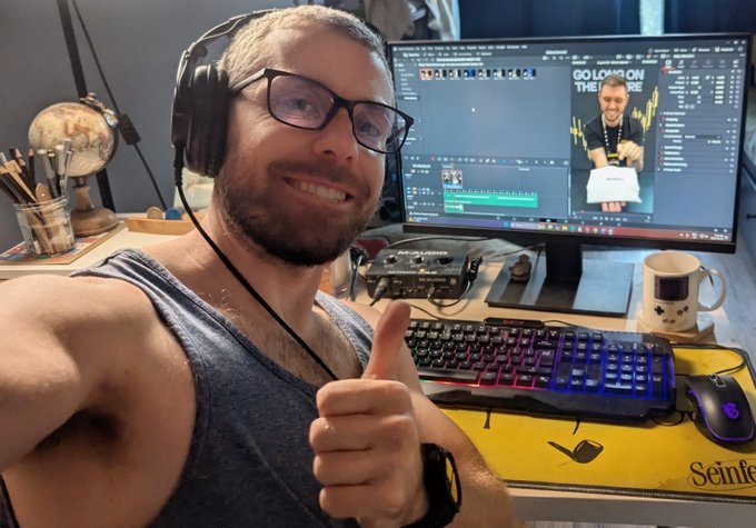 A smiling man with glasses and headphones gives a thumbs-up in front of a desk setup featuring a gaming keyboard, mouse, globe, and a monitor displaying video editing software with a 'GO LONG ON THE FUTURE' graphic visible on screen.