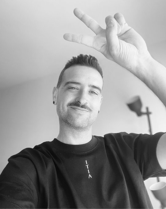 A man with a mustache smiles while holding up his hand in a gesture above his head, wearing a black shirt with "T T K" printed on the chest. The relaxed, celebratory pose pairs with the "GM big Fam, New week, WAGMI" message to convey an upbeat, community-focused greeting.