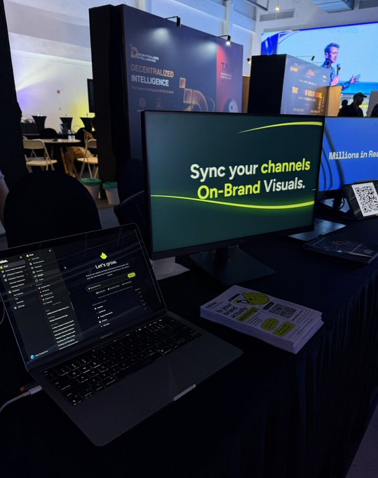 A convention display booth showing a laptop, a monitor displaying "Sync your channels On-Brand Visuals," and a background banner reading "DECENTRALIZED INTELLIGENCE" with a "7" logo.