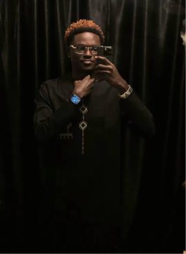 A man with orange-tinted hair wearing glasses and a black shirt, taking a mirror selfie with his phone in a dimly lit room with black curtains.