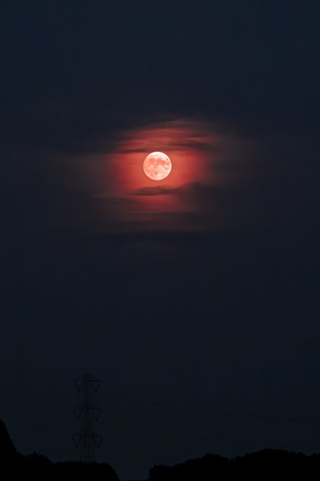A large, glowing red full moon sits in a dark, cloudy sky above the silhouette of a power line tower and hilly terrain.