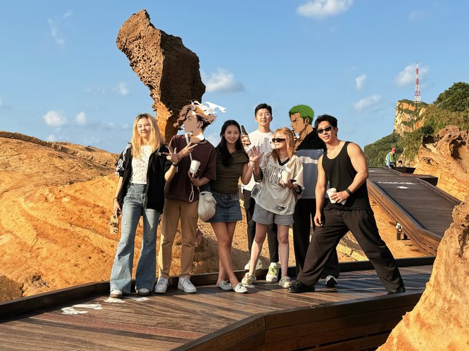 A group photo of seven people posing on a wooden path in a rocky, outdoor landscape with a large rock formation in the background.