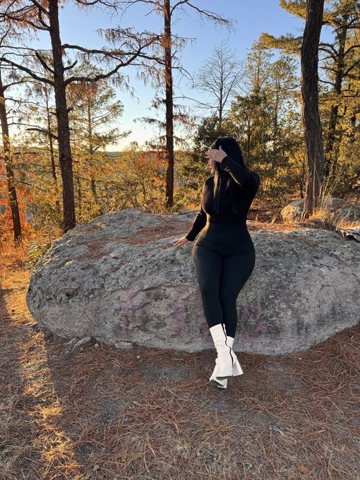 A person dressed in black athletic wear and white boots posing on a large boulder in a wooded area with autumn foliage and pine trees.