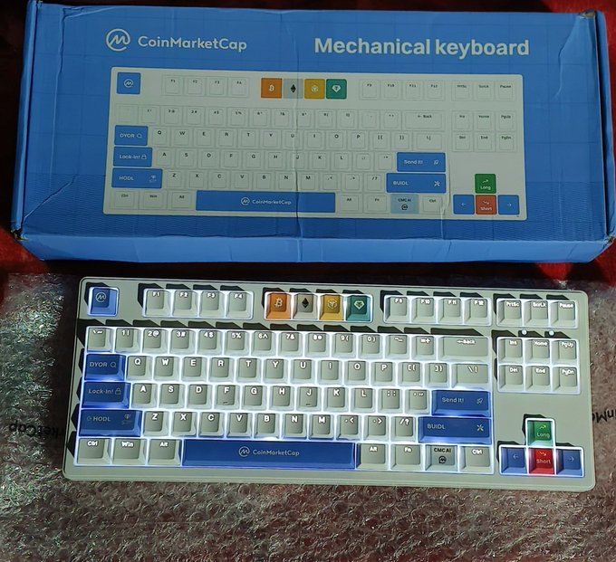 A CoinMarketCap branded mechanical keyboard is displayed in front of its blue packaging, which is labeled "CoinMarketCap Mechanical keyboard."