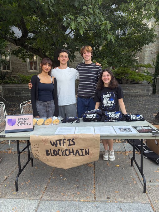 Four young people standing behind a table at an outdoor event with a "WTF IS BLOCKCHAIN" sign, "Stand With Crypto" materials, cookies, and "Build the future" merchandise.