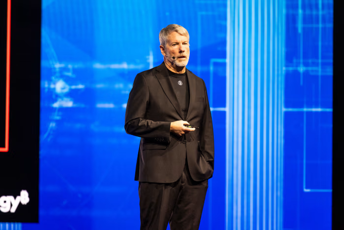 Michael Saylor, executive chairman of MicroStrategy and prominent Bitcoin advocate, speaks at a conference stage with a blue digital backdrop, visually tying to the news about his call for Brazil's first Bitcoin treasury ETF, which utilizes his company's $STRC and $SATA to deliver monthly dividends.