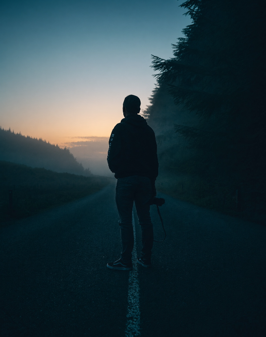 A silhouette of a person wearing a hoodie standing on a foggy road at sunset, with dark trees lining the side and holding what appears to be a camera.