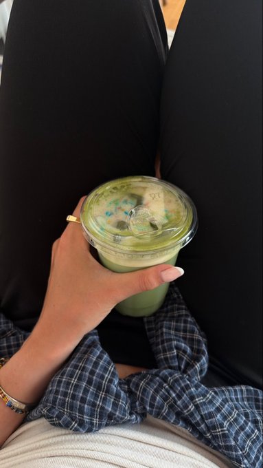 A hand holds a plastic cup of iced green matcha latte between a person's legs in black leggings.
