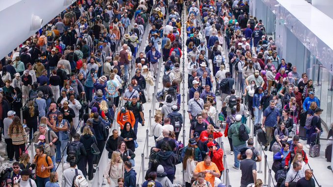 A dense crowd of travelers fills an airport terminal, waiting in long, snaking lines separated by stanchions, with luggage carts and passengers of diverse appearances visible throughout. The image illustrates the chaotic, overwhelming nature of crowded spaces—mirroring the "2026 in web3" theme of navigating a bustling, congested digital landscape where users must patiently work through overwhelming volumes of activity and traffic.