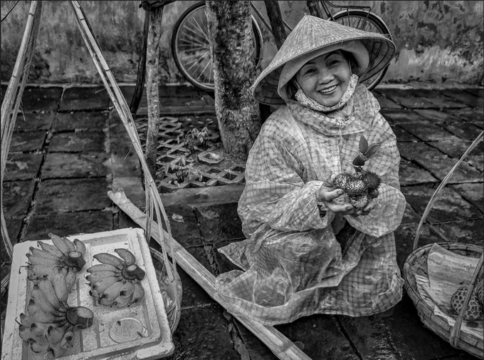 A black-and-white photo of a smiling woman wearing a conical hat, seated on the ground holding fruits, with a basket of bananas and a bicycle in the background.