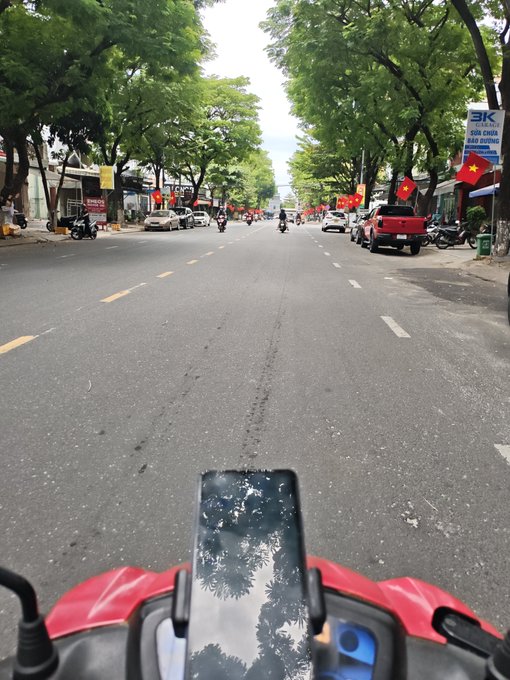 A first-person view from a scooter on a tree-lined urban street, with a smartphone mounted on the handlebars reflecting the trees and sky, and Vietnamese flags visible along the road.