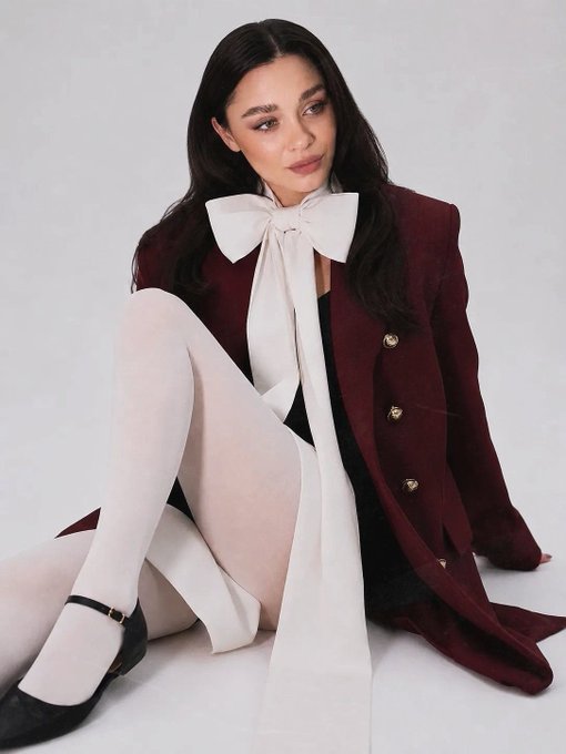 A woman with long dark hair sits elegantly in a burgundy double-breasted blazer, white tights, and black Mary Jane heels, styled with an oversized cream bow at the neckline. The minimalist studio portrait embodies the "In harmony with oneself" concept shared on the Tezos NFT marketplace objkt.com.